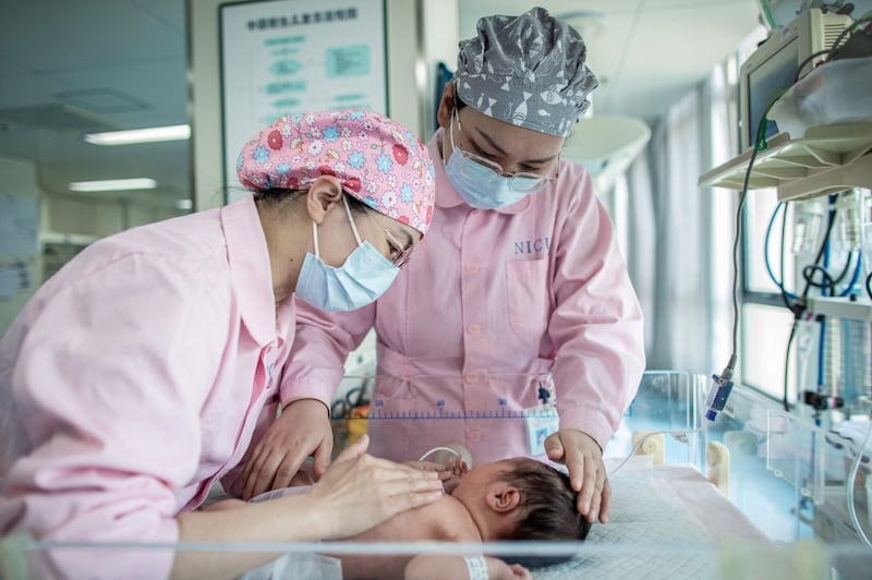 Two nurses wearing light pink scrubs and face masks lean over a newborn baby in a crib.